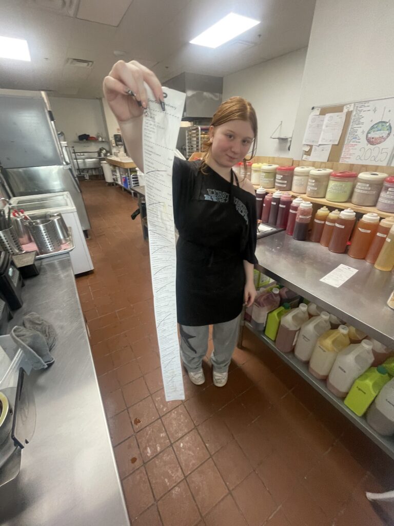 Violet at her job holding a long receipt in a restaurant kitchen.